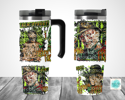 A travel mug shown from multiple angles on a gray wood floor against a white wall. The mug has a black handle and lid, with a wraparound horror-themed graphic featuring a zombie face, green graffiti-style splatters, and distressed text.