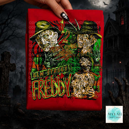 A hand holds a red graphic T-shirt with a text in green and yellow. The background is a dark, spooky graveyard scene with a haunted house.
