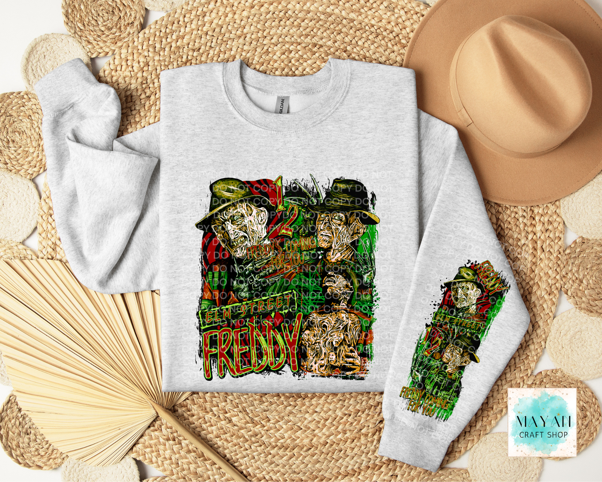 A light gray crewneck sweatshirt laid flat on a woven straw mat, featuring a large multicolor graphic on the front and a smaller matching graphic on the sleeve. A tan hat, folded fan, and woven mat surround the sweatshirt.