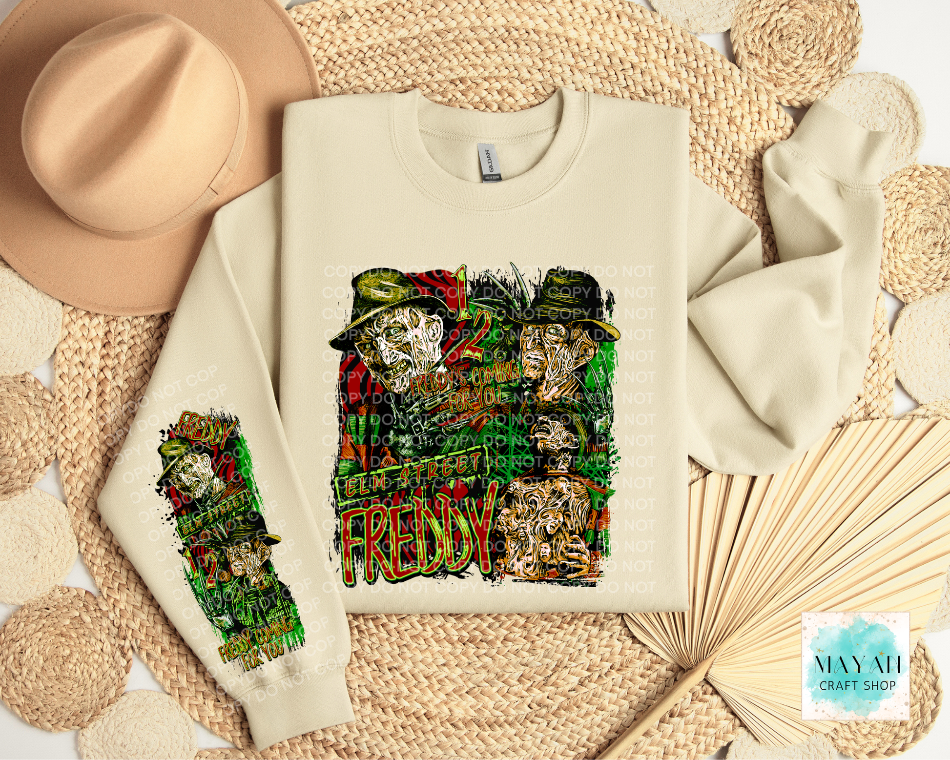 A cream-colored crewneck sweatshirt featuring a detailed graphic print. The sweatshirt is displayed in a styled flat-lay arrangement with a beige fedora hat, a woven mat, and a paper fan.