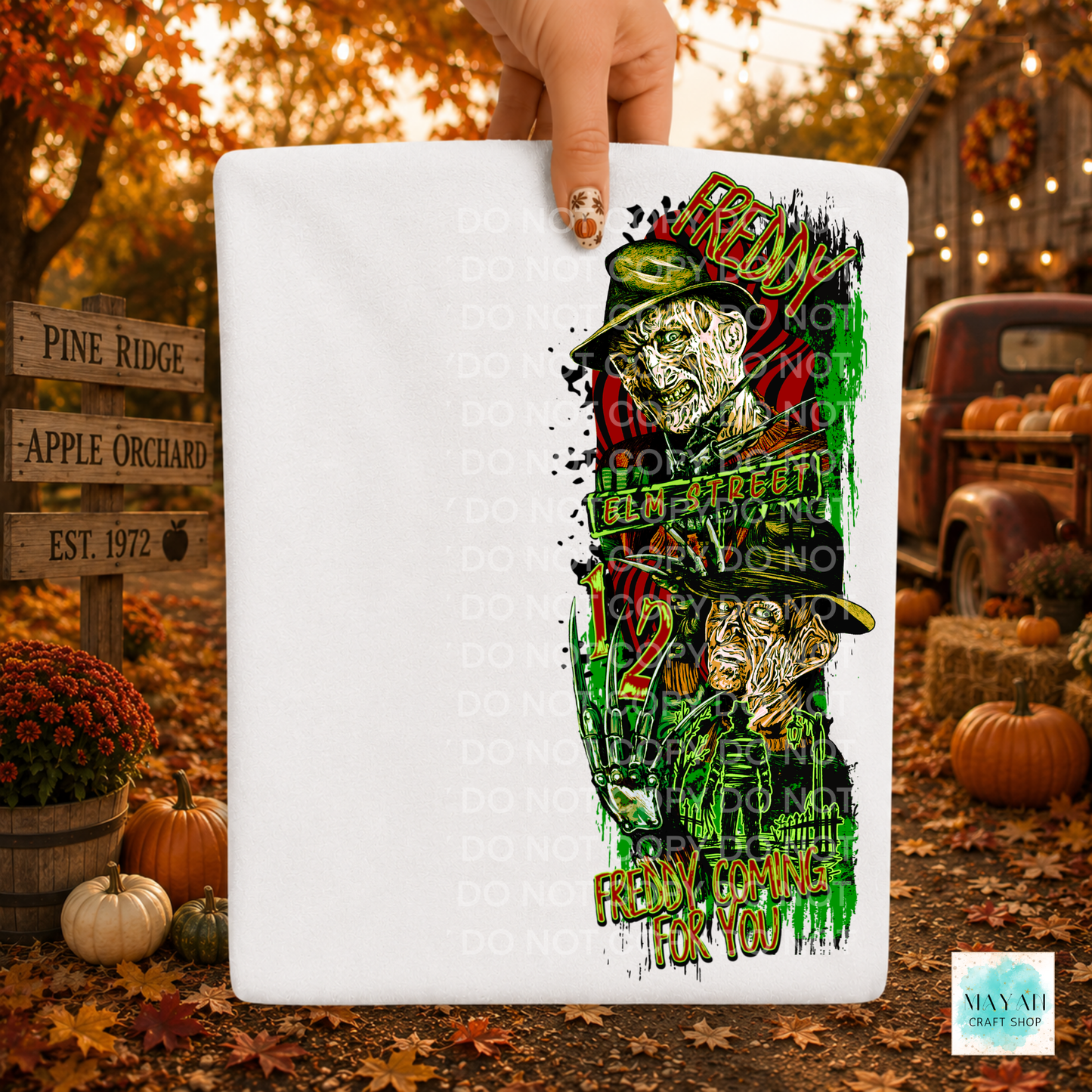 A white rectangular T-shirt or poster-like product held by a hand, featuring a colorful Halloween-style graphic with a horror character. The background is a fall yard scene with pumpkins, autumn leaves, and a rustic truck.