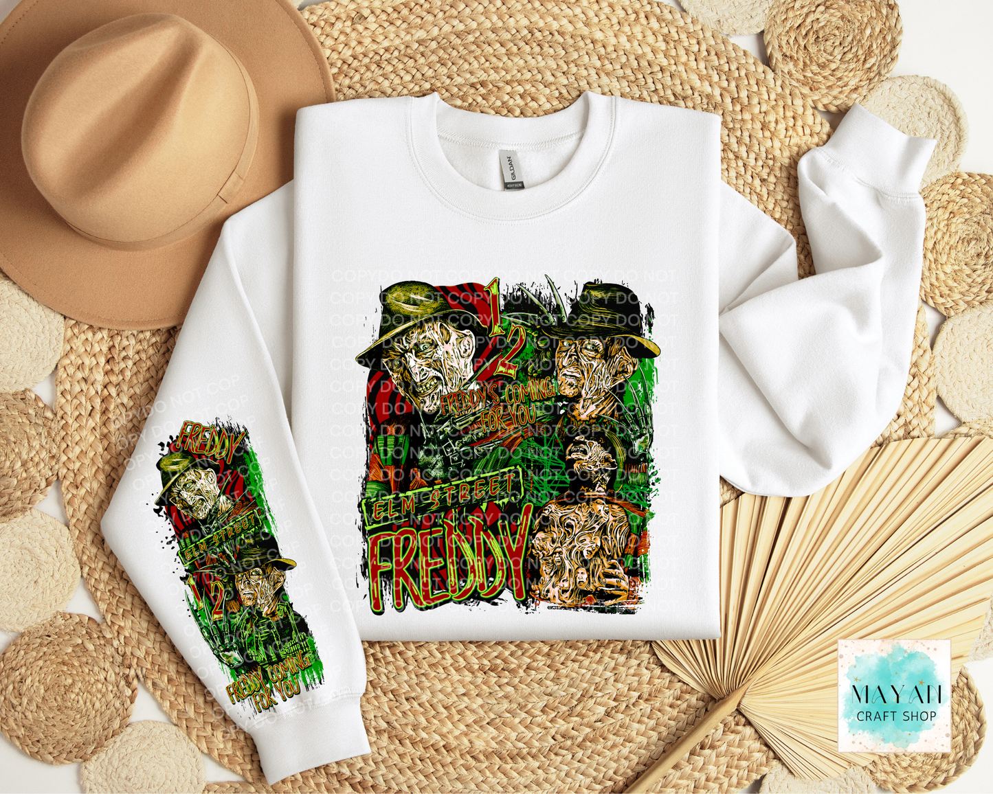 A white crewneck sweatshirt laid flat on a woven straw mat, featuring a large colorful graphic print on the front and a smaller matching sleeve print. A tan hat, woven mat, and dried palm leaves surround it.