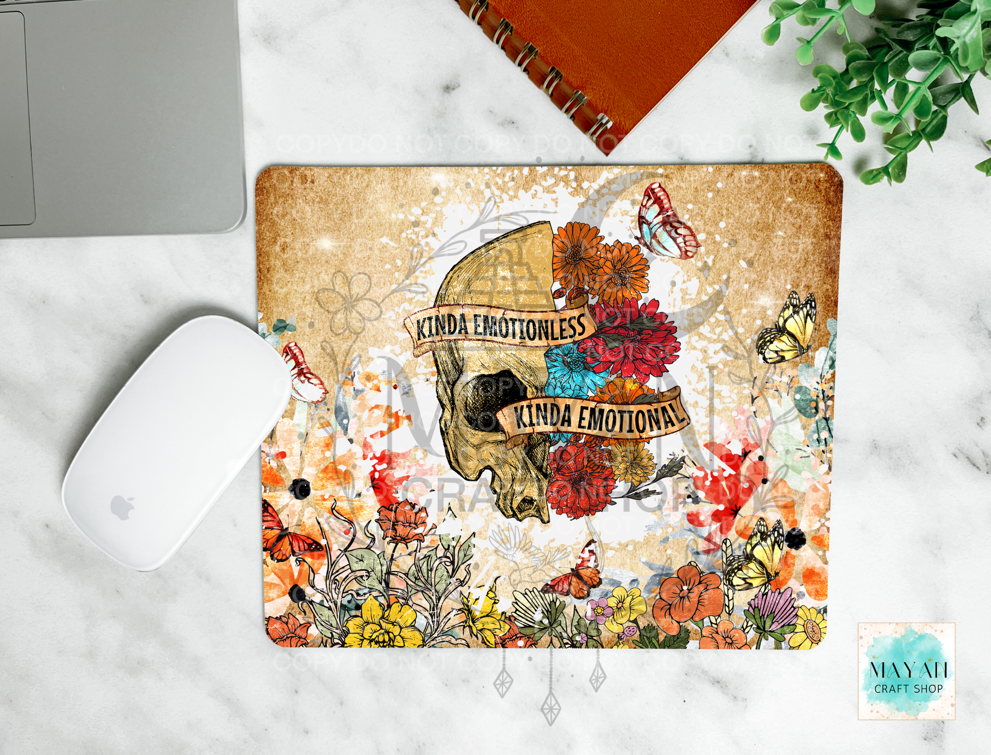 A mouse pad with a vintage-style skull and floral design sits on a white marble surface. The mat features the text 'KINDA EMOTIONLESS' and 'KINDA EMOTIONAL' over a skull surrounded by colorful flowers and butterflies.