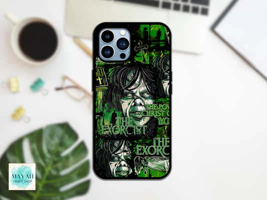 A black phone case with a green and black illustrated design featuring a text and stylized faces, shown on a white desk with a laptop, coffee cup, plant leaf, and binder clips in the background.