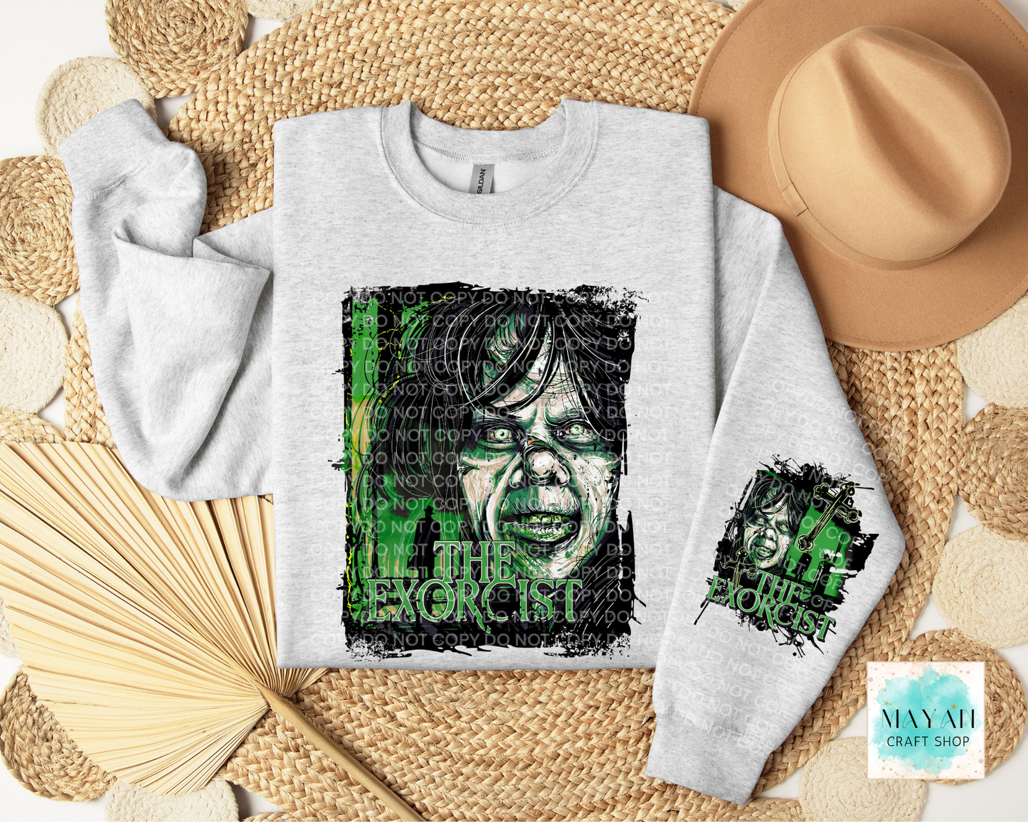 A light gray crewneck sweatshirt laid flat on a woven straw mat, featuring a large green and black graphic print of a horror character's face in the front, plus a smaller matching graphic on the left sleeve. A tan hat, folded straw fan, and woven mat surround the sweatshirt.