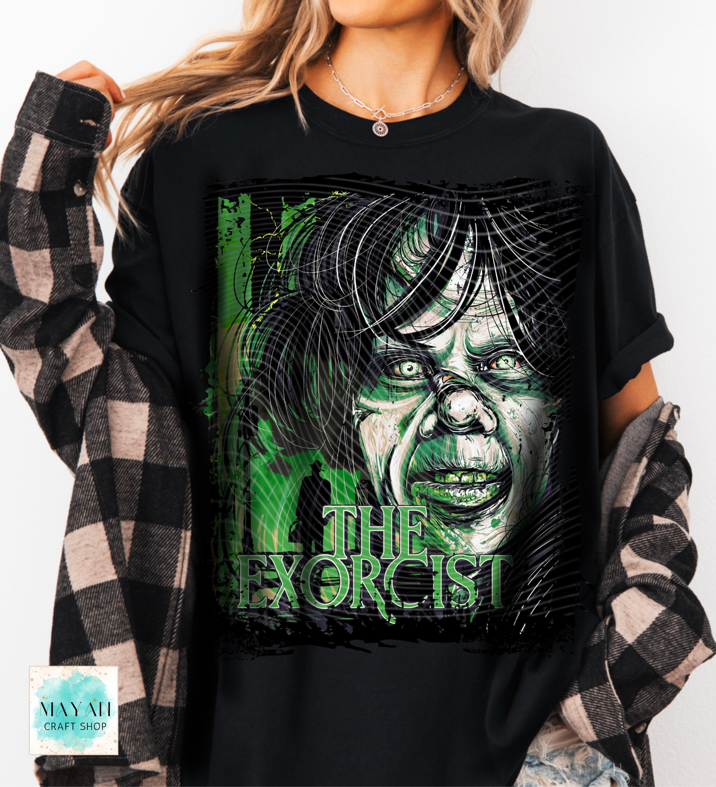 A woman wearing a black graphic t-shirt featuring a stylized green and white illustration of a character with the title printed below. She is wearing a black and white plaid flannel shirt as an overlay.