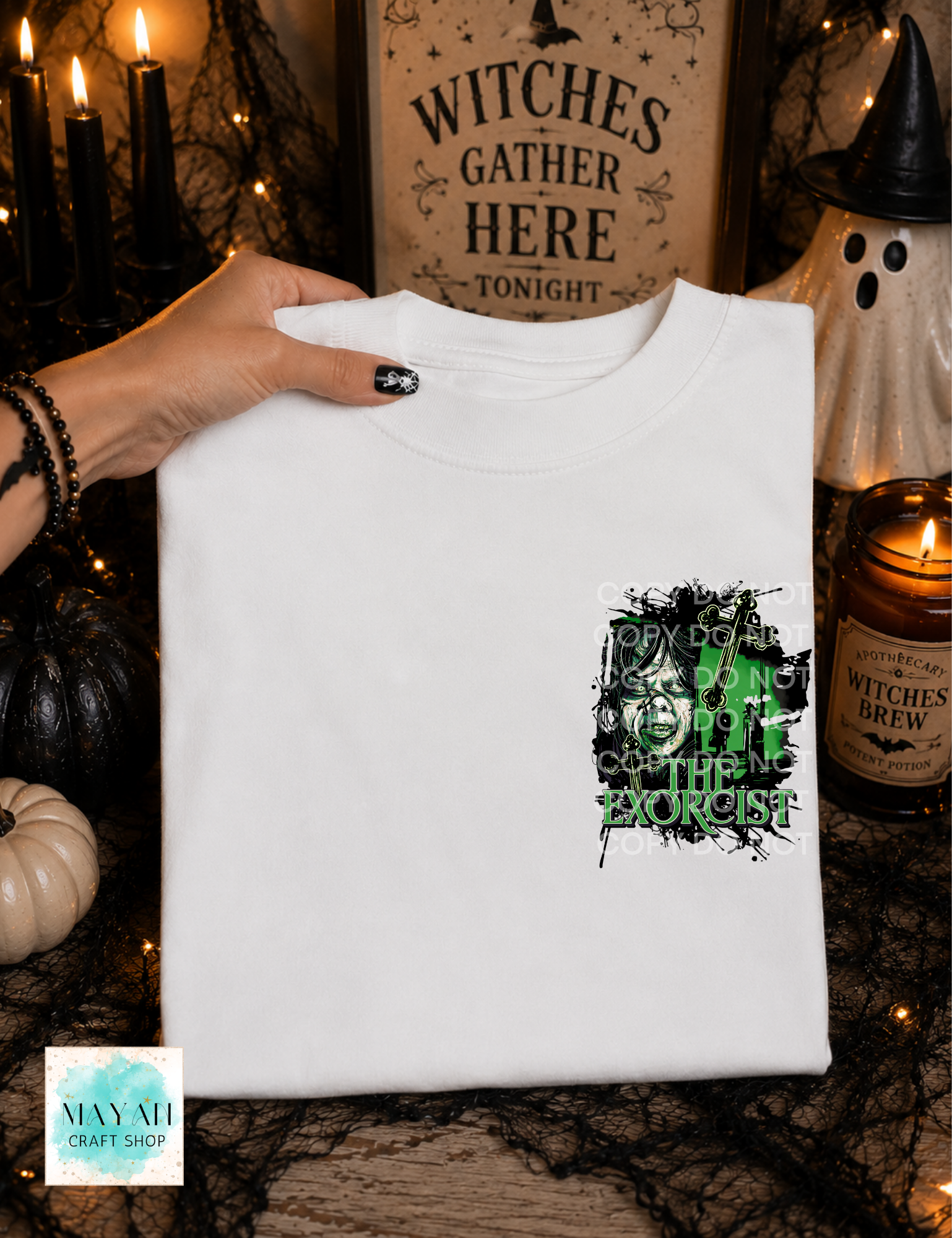A white crewneck t-shirt is held by a hand against a spooky Halloween-themed background. The shirt features a graphic on the left chest with a black background, a green witch hat.