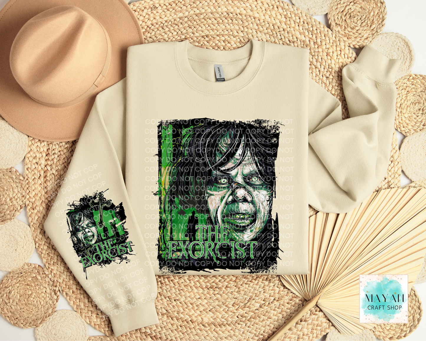 A cream-colored sweatshirt with a green and black graphic print of a face, styled in a flat lay with a tan hat, woven mat, and paper fan.