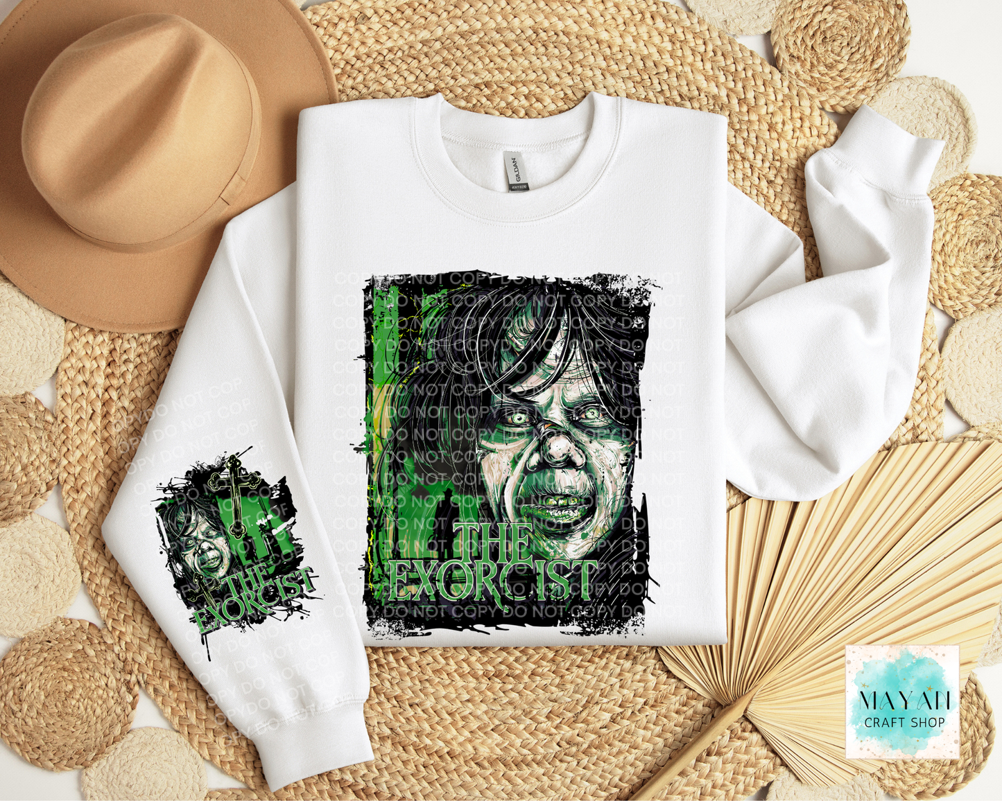 A white crewneck sweatshirt with a green and black graphic print of a face displayed in a flat-lay arrangement with a tan hat, woven mats, and a paper fan.