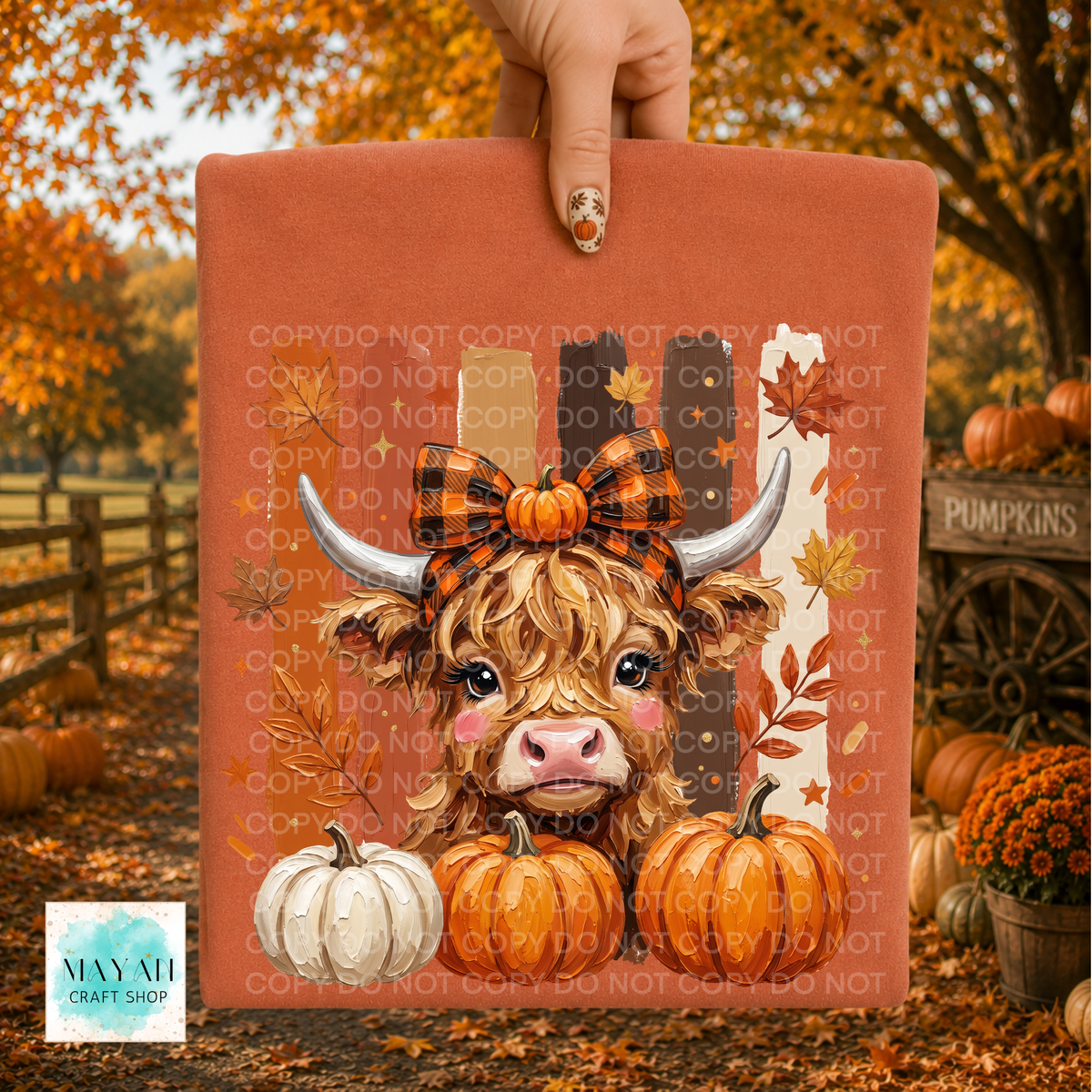 A hand holds a shirt featuring a cartoon Highland cow with a black-and-orange bow, white and orange pumpkins, and autumn leaves. The background is a blurred fall yard scene with pumpkins, orange foliage, and a wagon wheel.