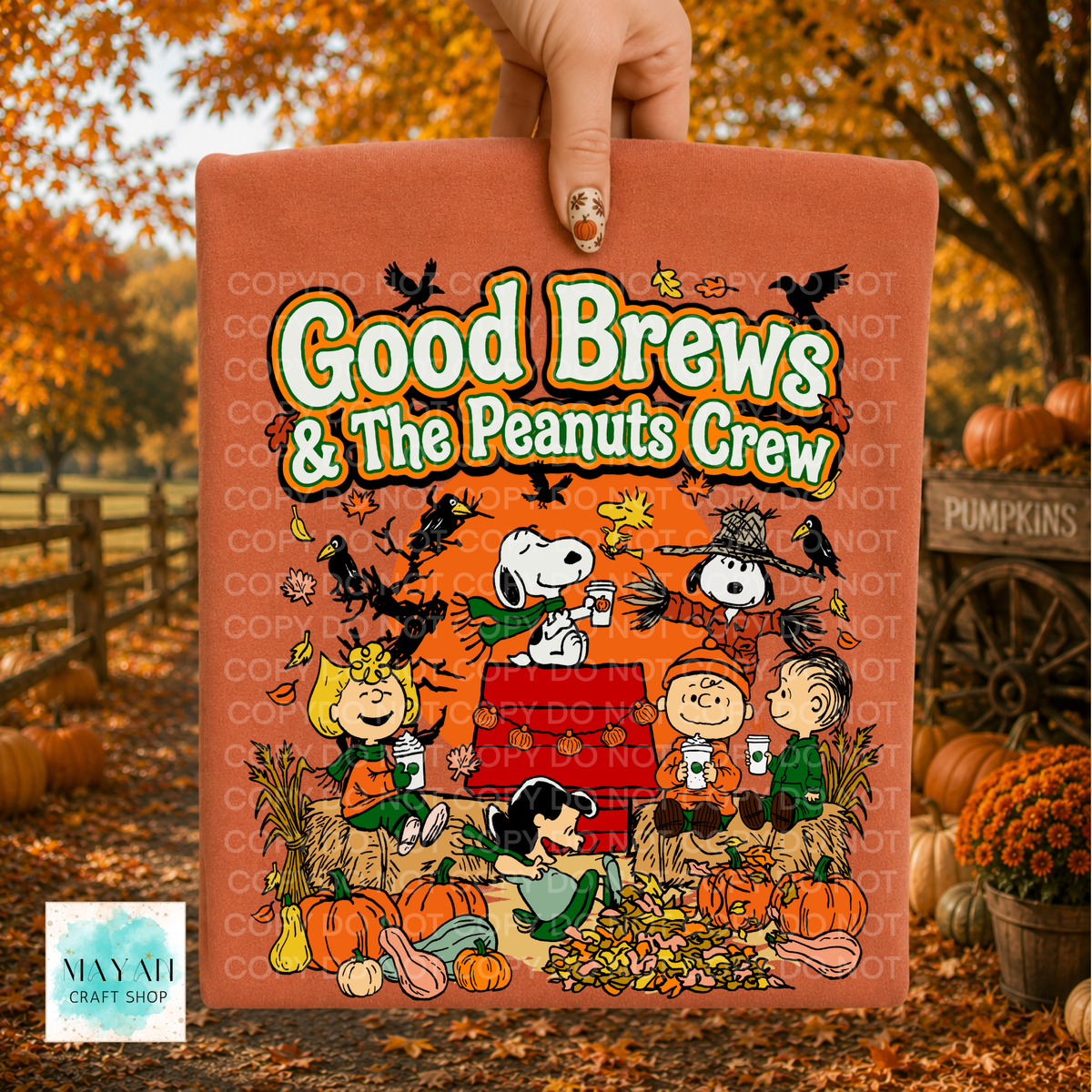 A hand holds a orange shirt featuring a colorful cartoon illustration of characters with the text 'Good Brews' The background shows an autumn outdoor scene with pumpkins, fall leaves, and a fence.