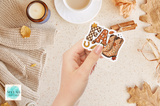 A hand holds a die-cut sticker featuring the word 'FALL' in a patterned font with autumn-themed embellishments like a horseshoe, leaf, and cinnamon stick, set against a cozy flat-lay background with a cup of coffee, candle, and leaves.