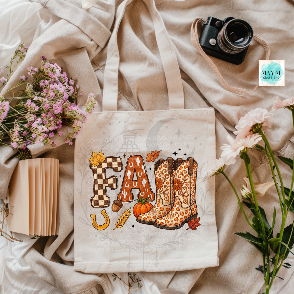A cream canvas tote bag with a fall-themed graphic reading 'FALL' in checkered and leopard-print letters, surrounded by autumn motifs like pumpkins, leaves, and a horseshoe. The bag is styled in a flat lay with flowers, a camera, and a notebook on a beige fabric background.