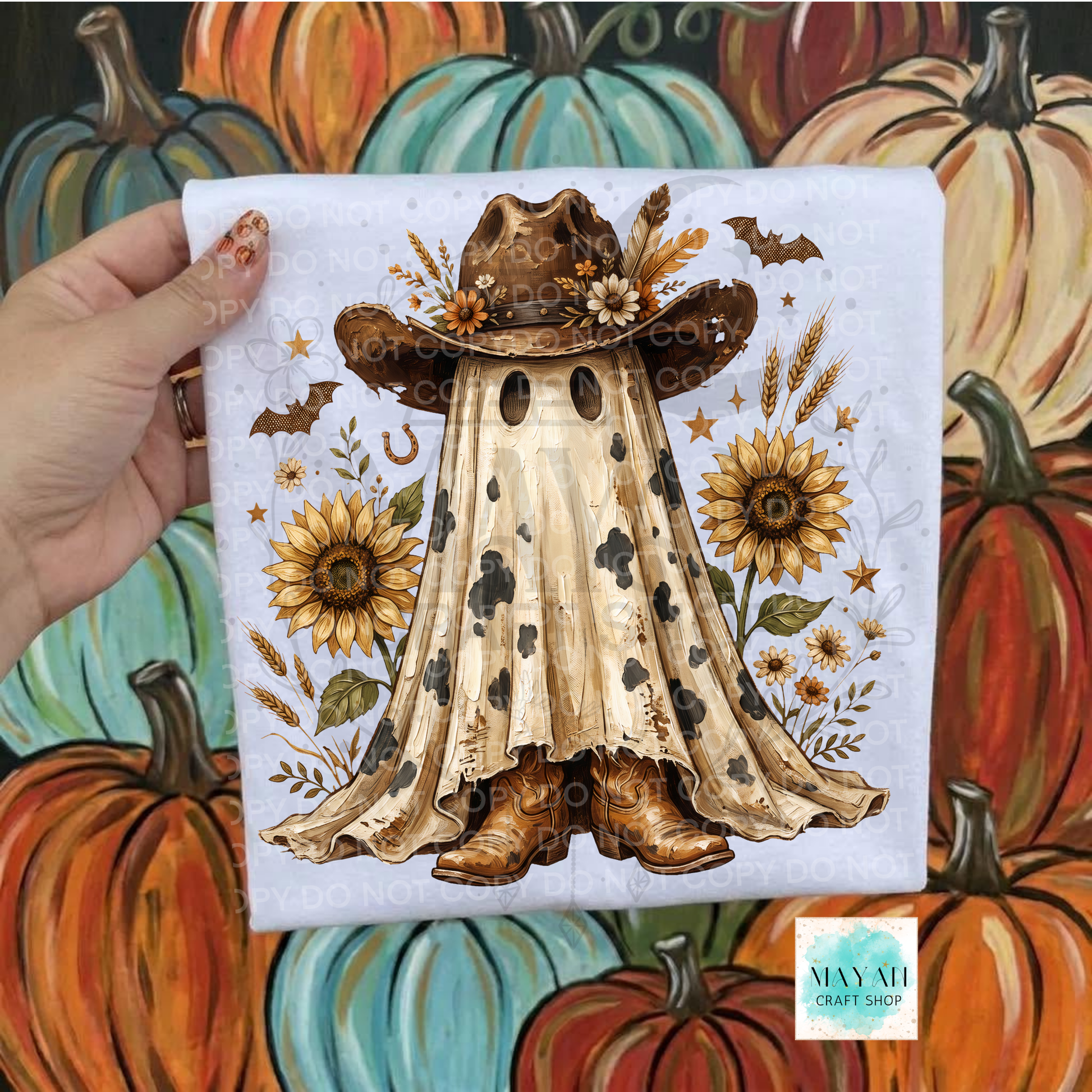 A hand holds a square white fabric print featuring a ghost-like cowboy figure in a hat and boots, surrounded by sunflowers, wheat, bats, and stars. The background is a painted autumn scene with pumpkins.