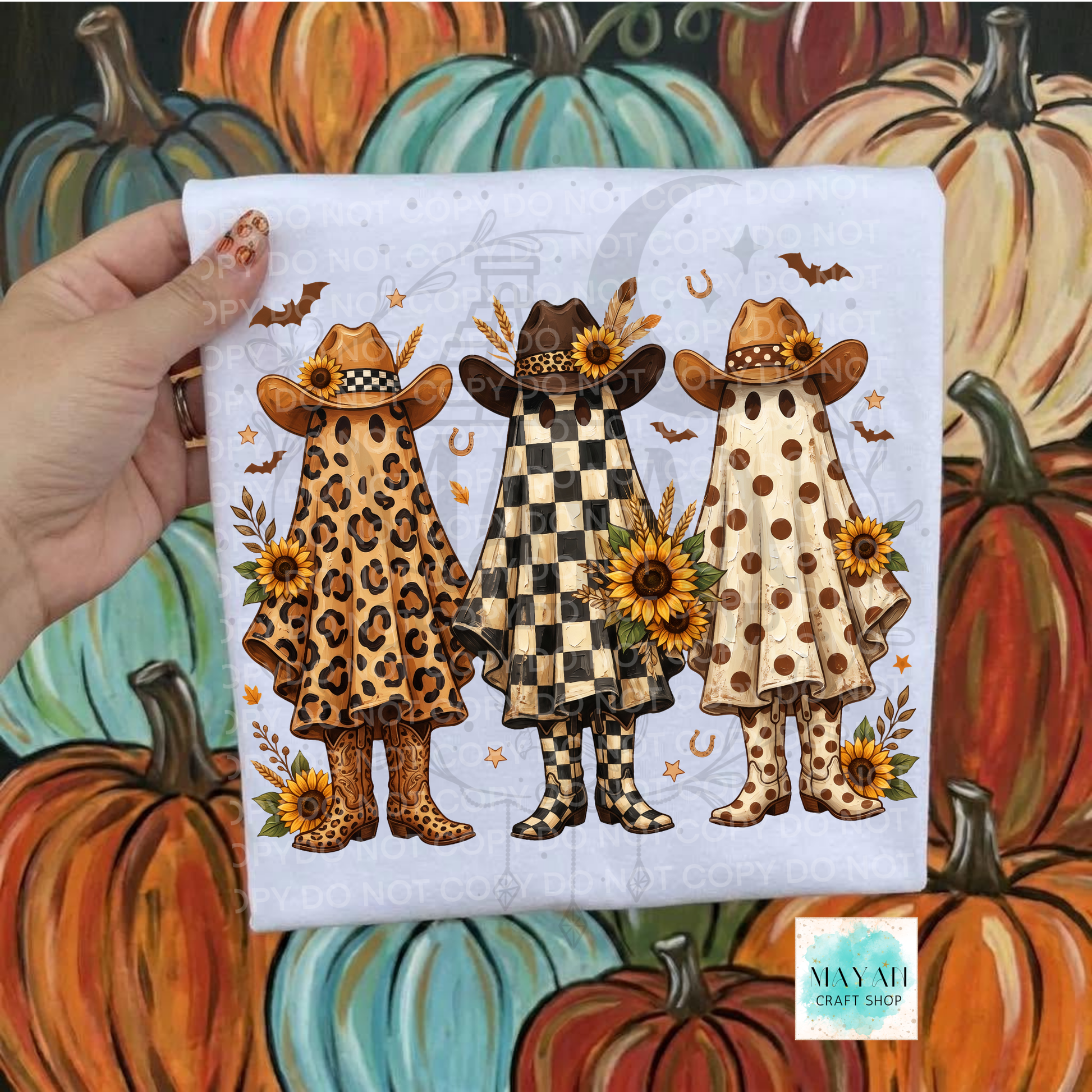 A hand holds a square white fabric print featuring three stylized ghosts in autumn-themed outfits: leopard print, black-and-white check, and beige with brown polka dots, decorated with sunflowers, bats, horseshoes, and stars. The background is a painted pumpkin scene.