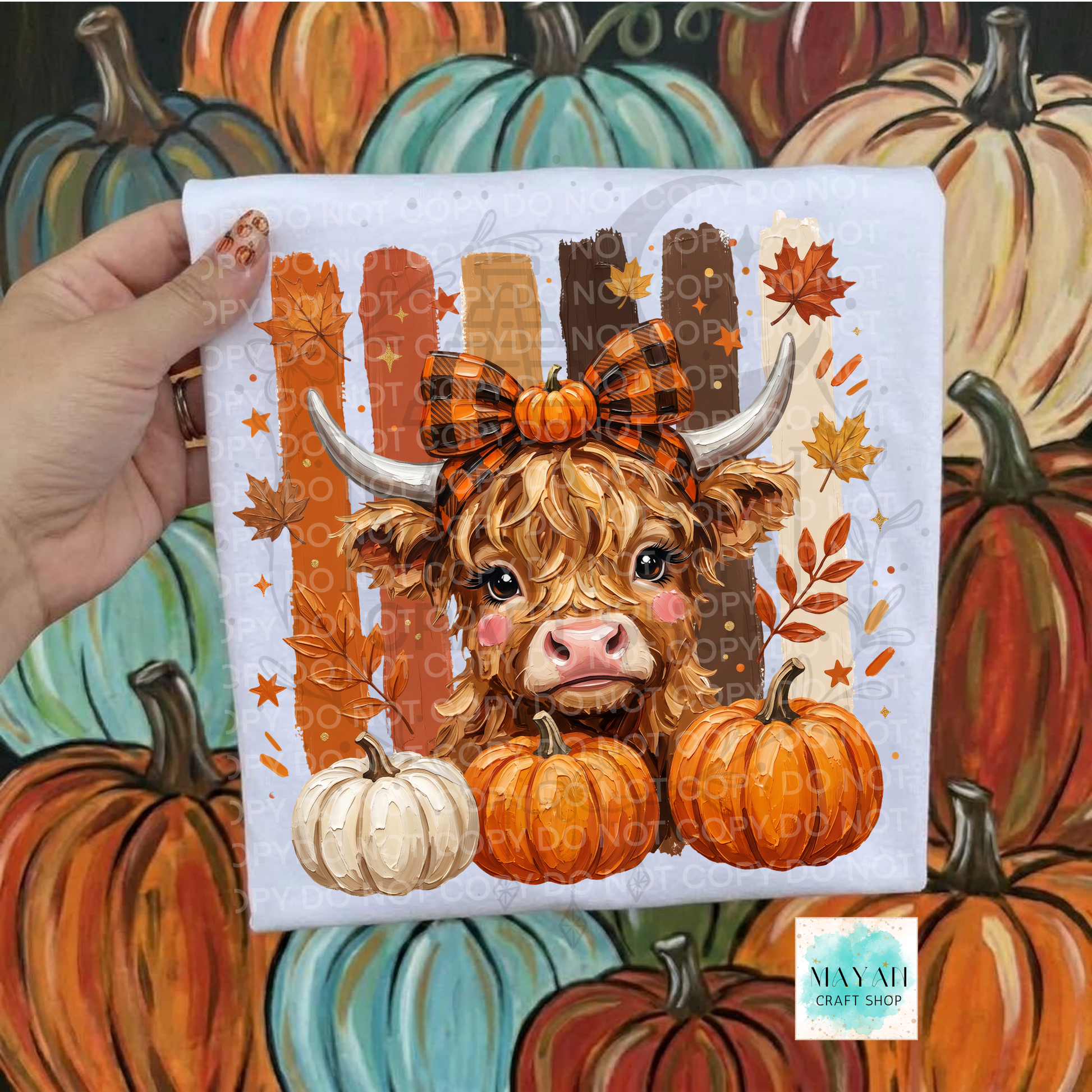 A hand holds a square fabric print featuring a cute Highland cow with a plaid bow, pumpkins, and autumn leaves. The background is a painted scene of pumpkins.