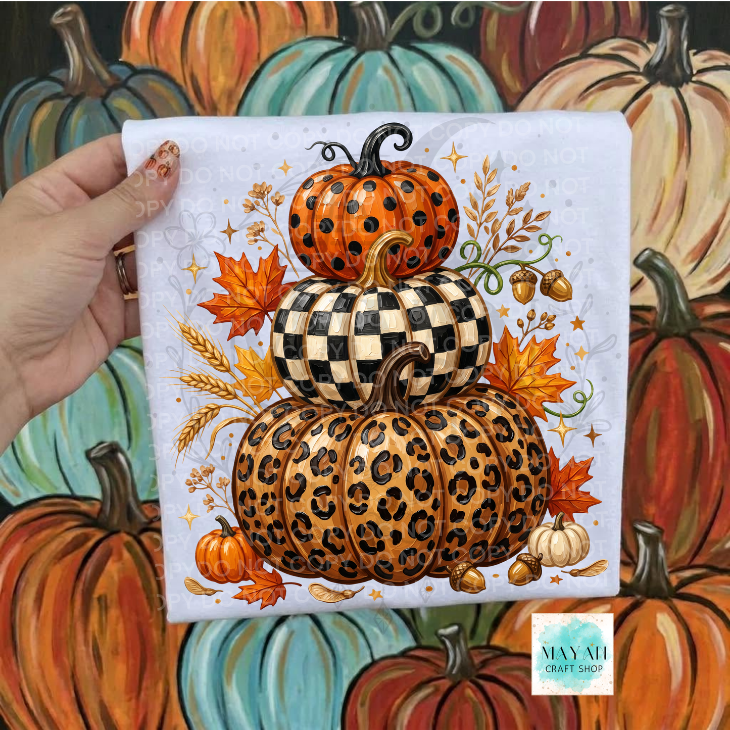 A hand holds a shirt featuring stacked pumpkins in orange, black-and-white checkered, and leopard print designs, surrounded by autumn leaves and acorns. The background is a painted scene of colorful pumpkins.
