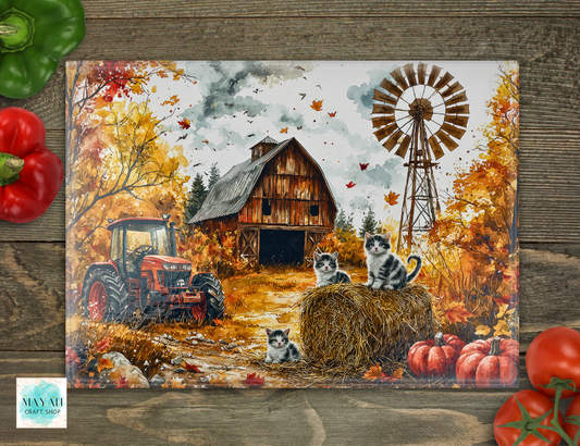 A cutting board featuring a fall farm scene with a barn, windmill, tractor, pumpkins, and several kittens, displayed on a rustic wood surface with decorative red peppers and tomatoes around it.