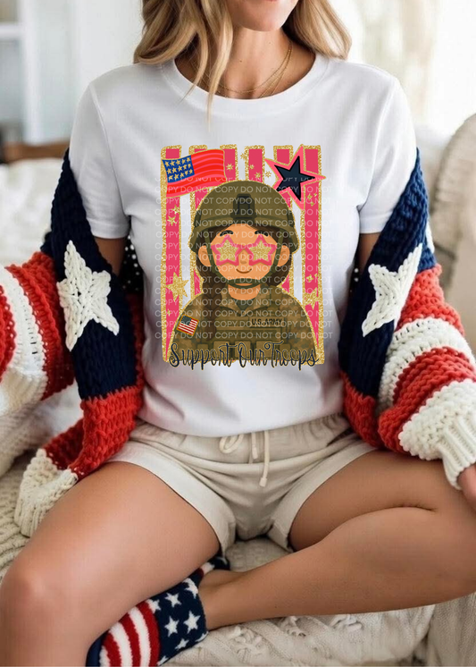 A woman sitting indoors wearing a white graphic T-shirt with a patriotic military-themed print, a red, white, and blue knit cardigan, light beige shorts, and matching socks.