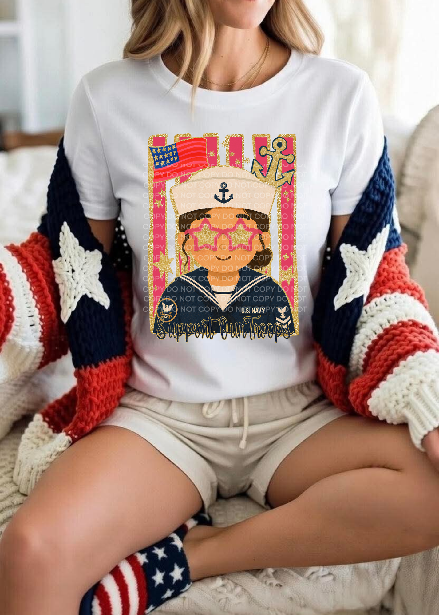 A woman sitting indoors wearing a white graphic T-shirt with a colorful patriotic sailor-themed print, a red, white, and blue knit cardigan, and beige shorts.