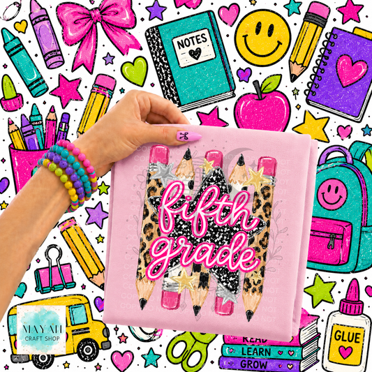 A hand holds a pink shirt featuring three illustrated pencils with leopard print, black glitter, and pink erasers, with the words 'fifth grade' in pink script. The background is a busy school-themed illustration with crayons, notebooks, stars, an apple, a backpack, and other stationery items.