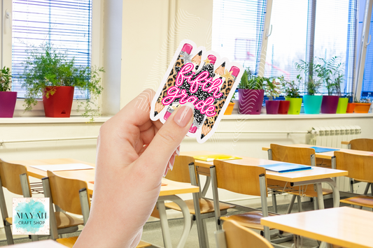 A hand holds a die-cut sticker featuring pink glitter-style text over a pencil illustration with leopard print and pink nail tips, set against a blurred classroom background with desks and potted plants.