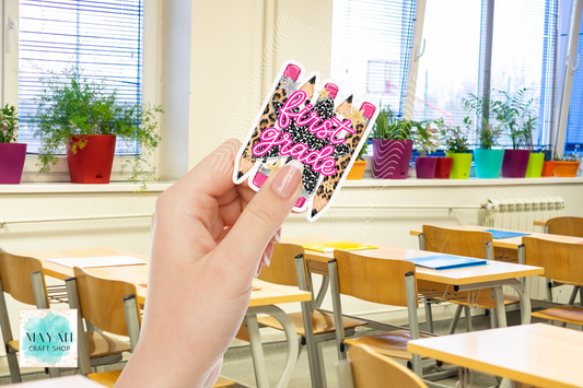 A hand holds a die-cut sticker featuring three pink and black leopard-print pencils, set against a blurred classroom background with desks and potted plants.