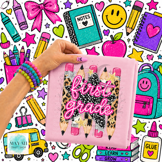 A hand holds a pink shirt featuring three illustrated pencils with leopard print, black glitter, and pink glitter tips, with the words 'first grade' in pink glitter script. The background is a busy school-themed graphic with crayons, notebooks, an apple, smiley face, backpack, and other stationery items.