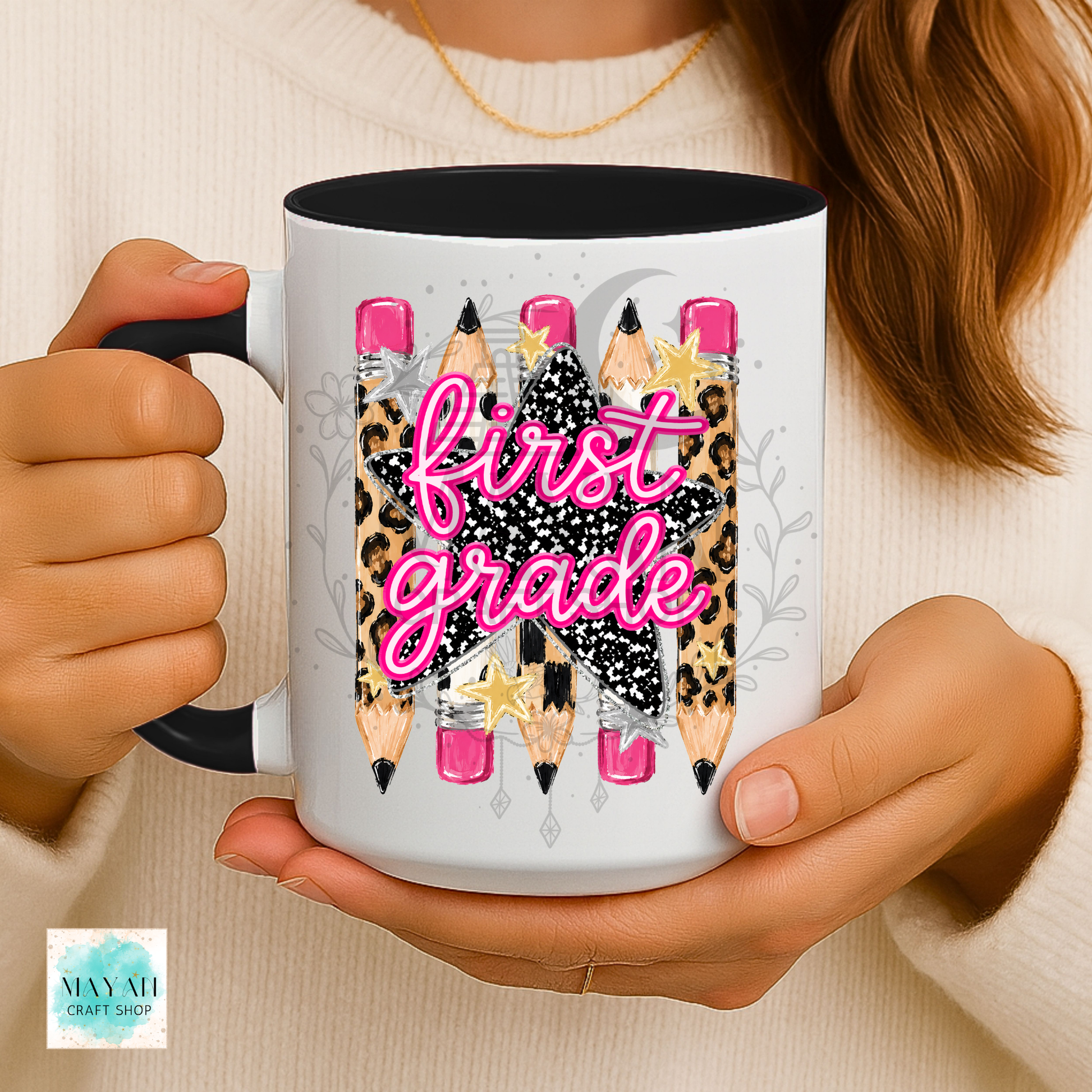 A person holds a white ceramic mug with a black handle and interior. The mug features a graphic of pencils with leopard print, pink erasers, and a black star with the words 'first grade' in pink script.