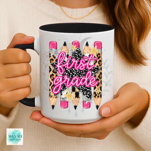 A person holds a white ceramic mug with a black handle and interior. The mug features a graphic of pencils with leopard print, pink erasers, and a black star with the words 'first grade' in pink script.