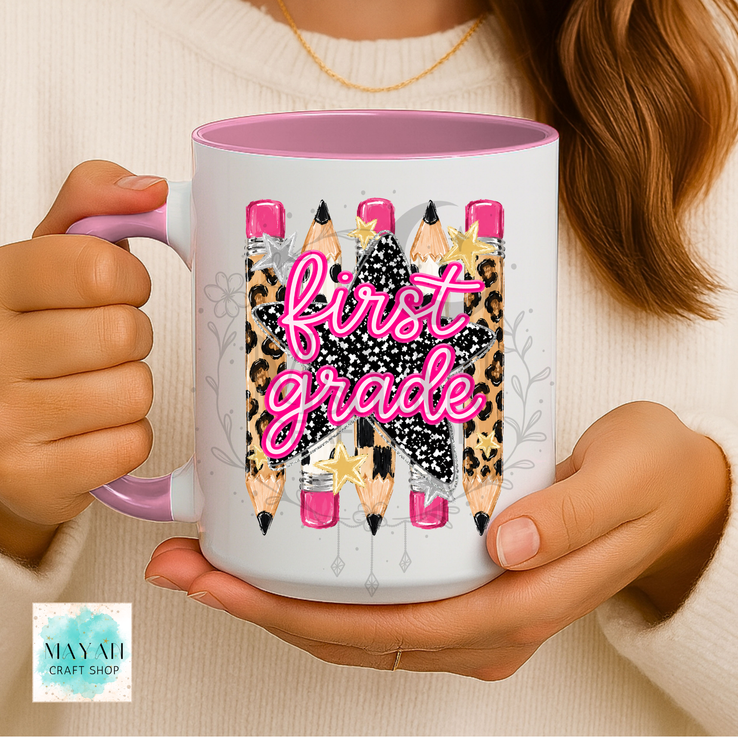 A person holds a white ceramic mug with a pink interior and handle. The mug features a graphic of five colored pencils with leopard print, black glitter, and gold stars, overlaid with the words 'first grade' in bright pink script.