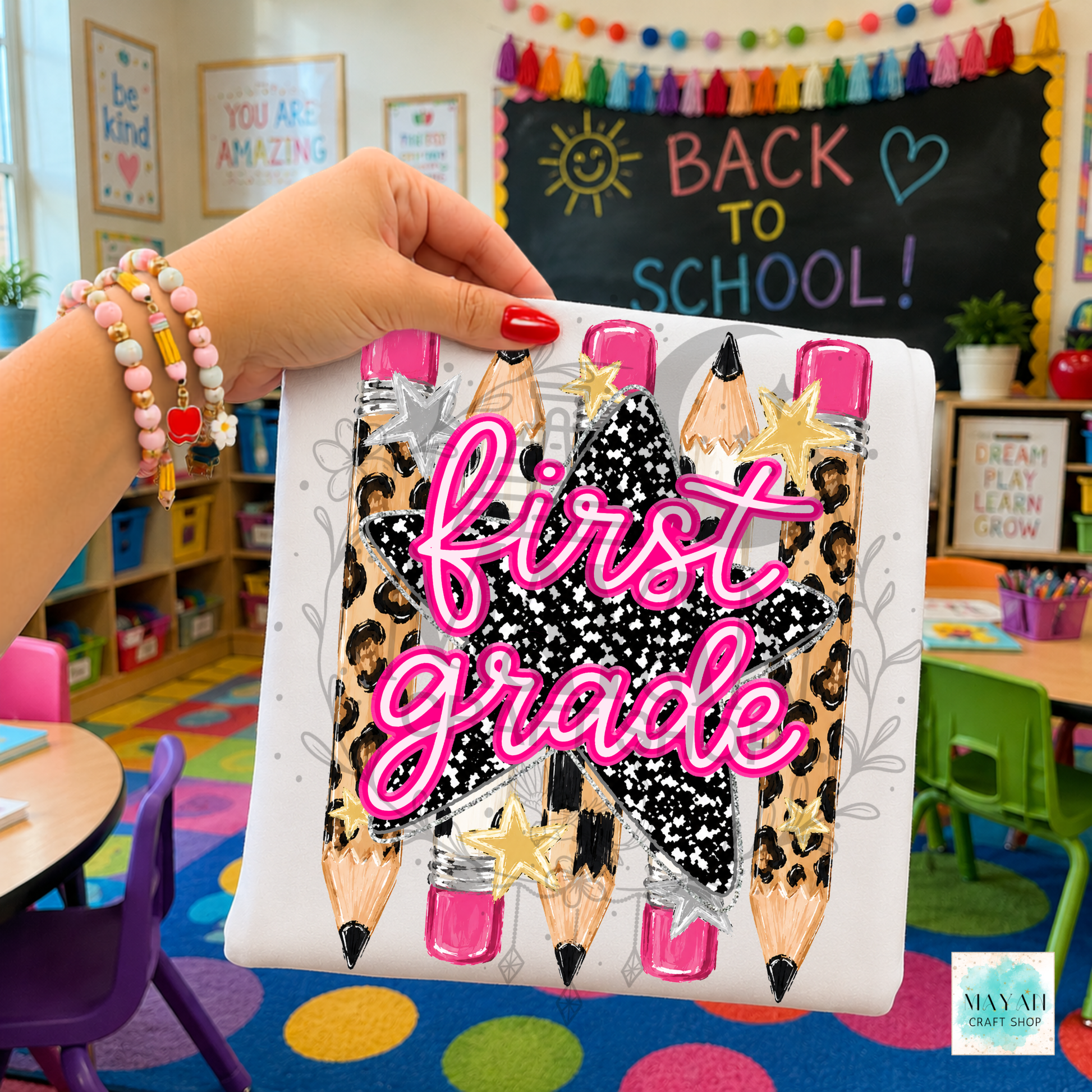 A hand holding a white shirt featuring a graphic design of three pencil illustrations with leopard print, black-and-white speckled centers, pink nail tips, and the words 'first grade' in bright pink script. The background shows a colorful children's classroom with a chalkboard that says 'BACK TO SCHOOL!'.