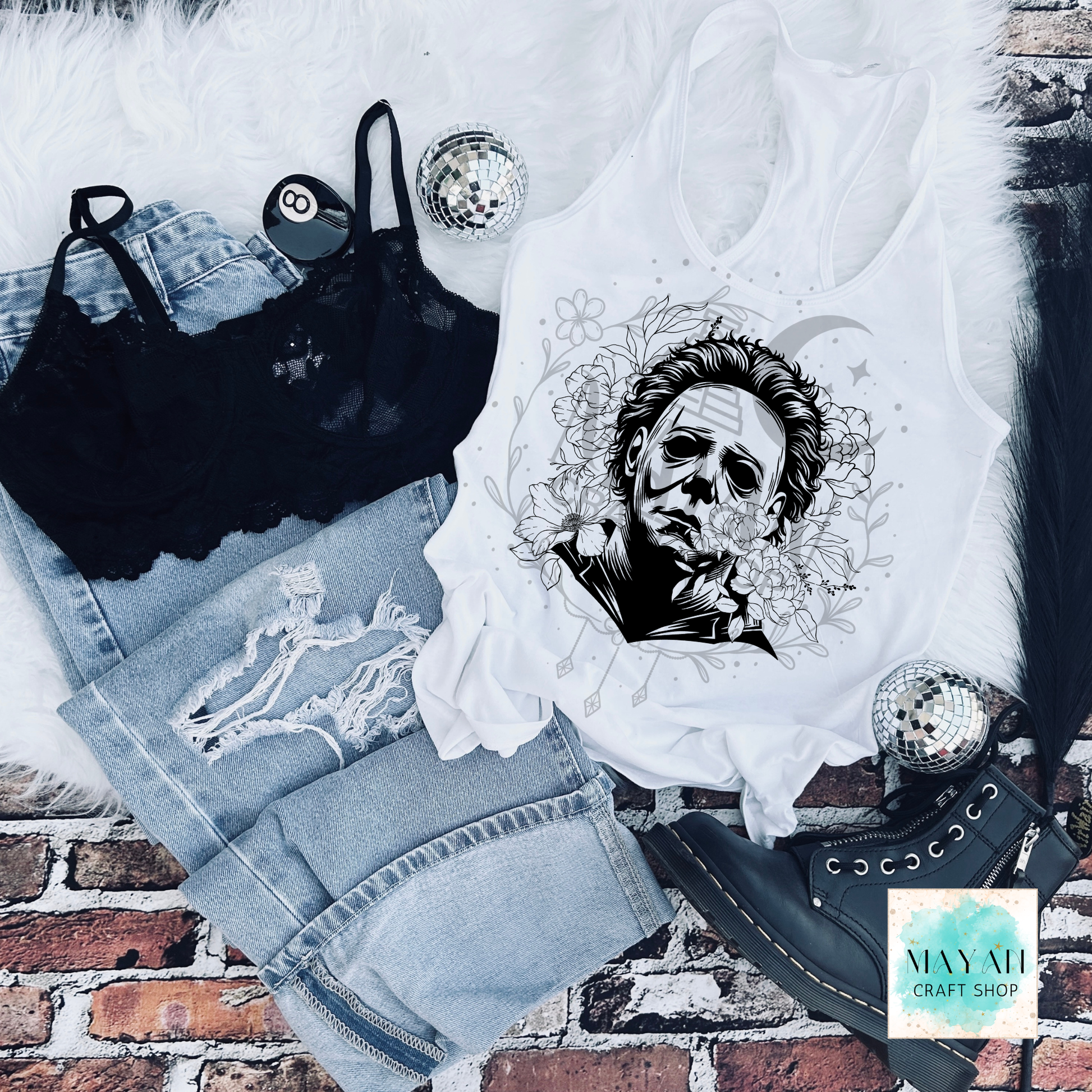 White tank top with black and white graphic design on a brick surface, surrounded by denim shorts and black lace items.