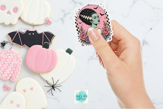 Decorative cookies shaped like a bat, pumpkin, and ghost with a hand holding a sticker of a green-skinned woman with black hair.