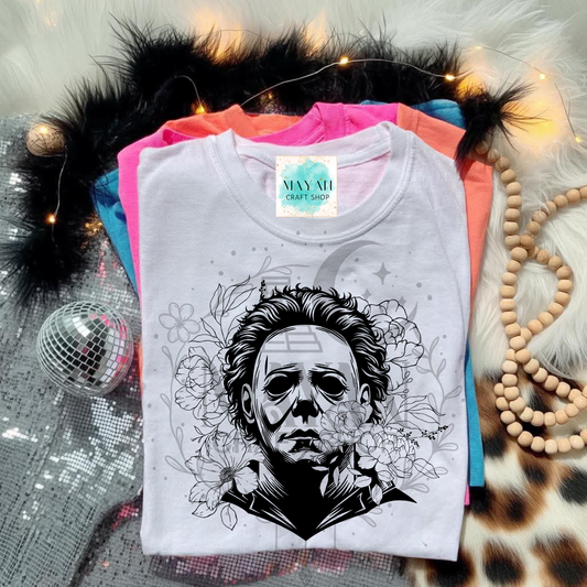 White t-shirt with black graphic design of a face and flowers, placed on colorful fabric with decorative items.