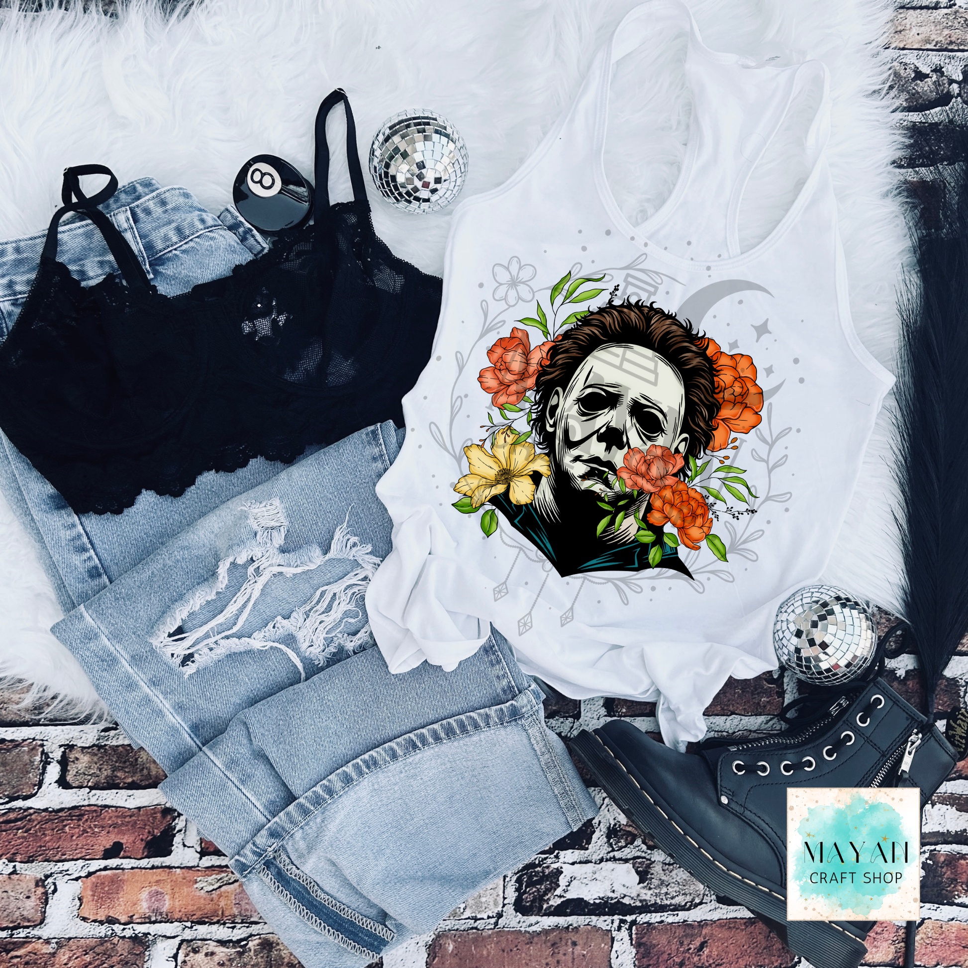 White tank top with a graphic design of a face surrounded by flowers, black bra, jeans, and black high-heeled shoes on a brick background.