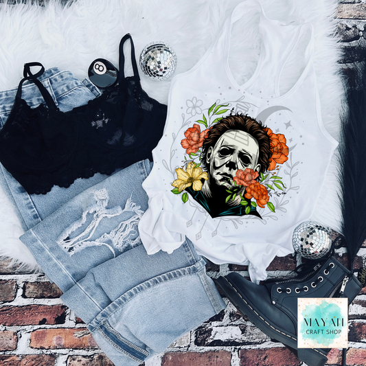 White tank top with a graphic design of a face surrounded by flowers, black bra, jeans, and black high-heeled shoes on a brick background.