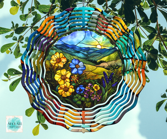 A wind spinner hanging with a colorful rainbow border and a scenic landscape insert of flowers, hills, and mountains. The piece is photographed hanging against a pale sky with green leaves around it.