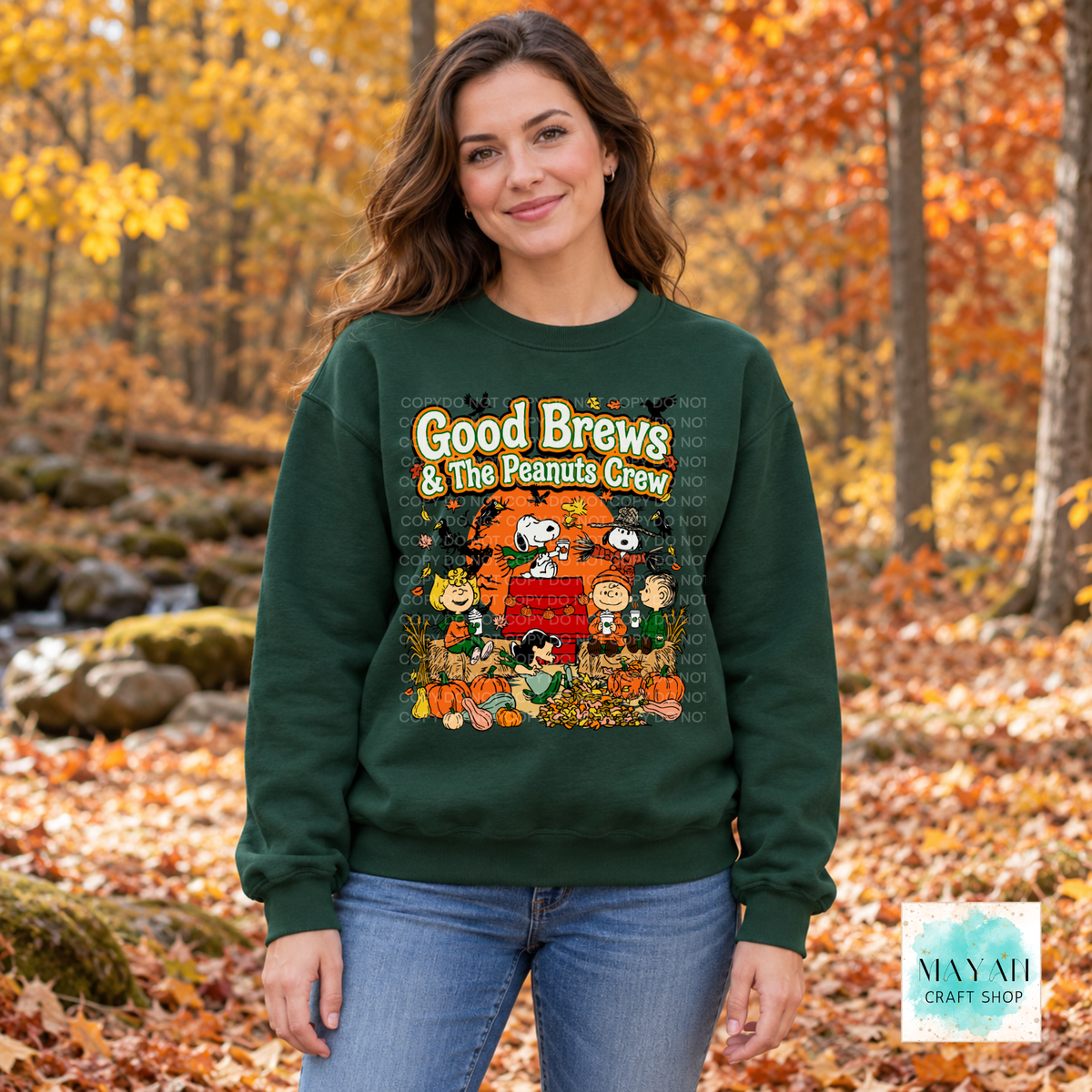 A woman wearing a dark green crewneck sweatshirt with a colorful 'Good Brews' graphic, standing in an autumn forest setting with fallen leaves.