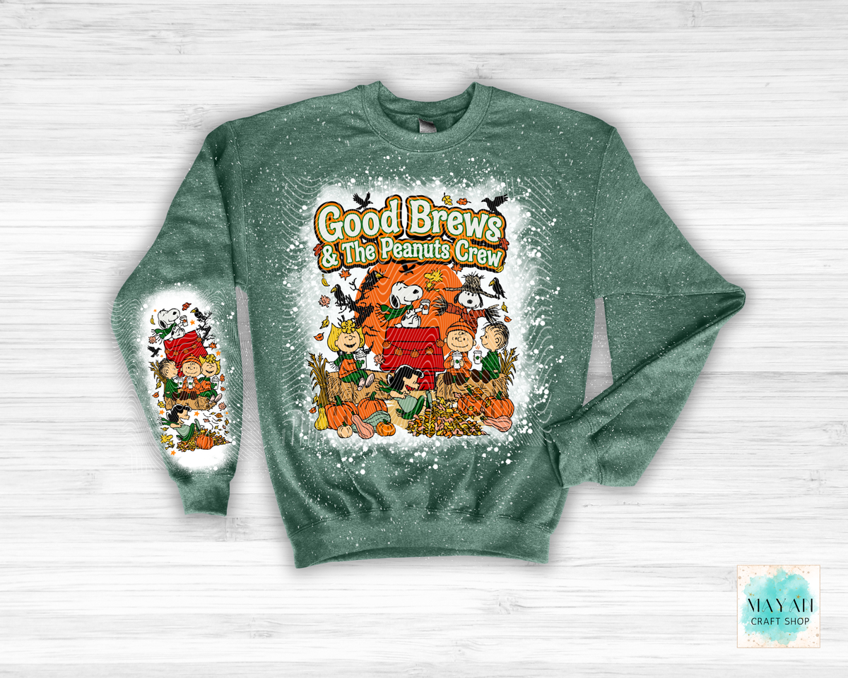 A heather green crewneck sweatshirt featuring a festive Fall graphic for 'Good Brews' with characters. The sweatshirt is displayed flat on a light gray wood-textured background, with a brand watermark in the bottom right corner.