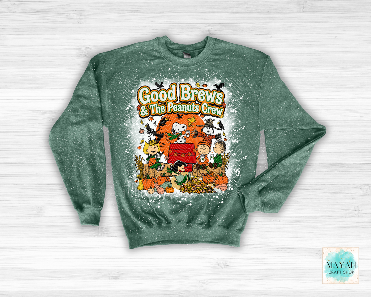 A heathered dark green crewneck sweatshirt featuring a festive 'Good Brews' graphic print with characters in a fall harvest scene, displayed flat on a white wood background.