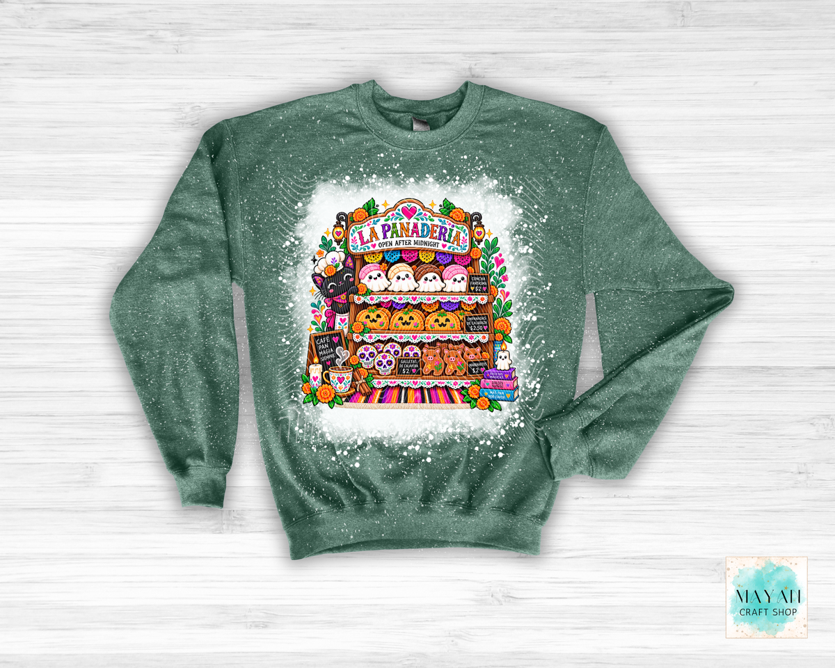 A green crewneck sweatshirt with a white distressed splatter effect, featuring a colorful graphic of a Mexican panaderia bakery stall with cute cartoon characters. The sweatshirt is laid flat on a white wood background.