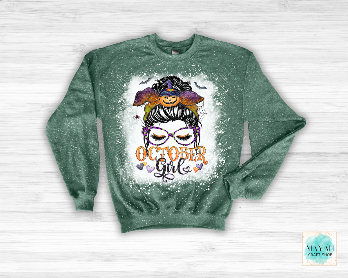 A green long-sleeve crewneck sweatshirt with a distressed splatter finish, featuring a graphic of a witchy girl with a pumpkin hat and the text 'OCTOBER Girl' on the front, displayed on a white wood background.