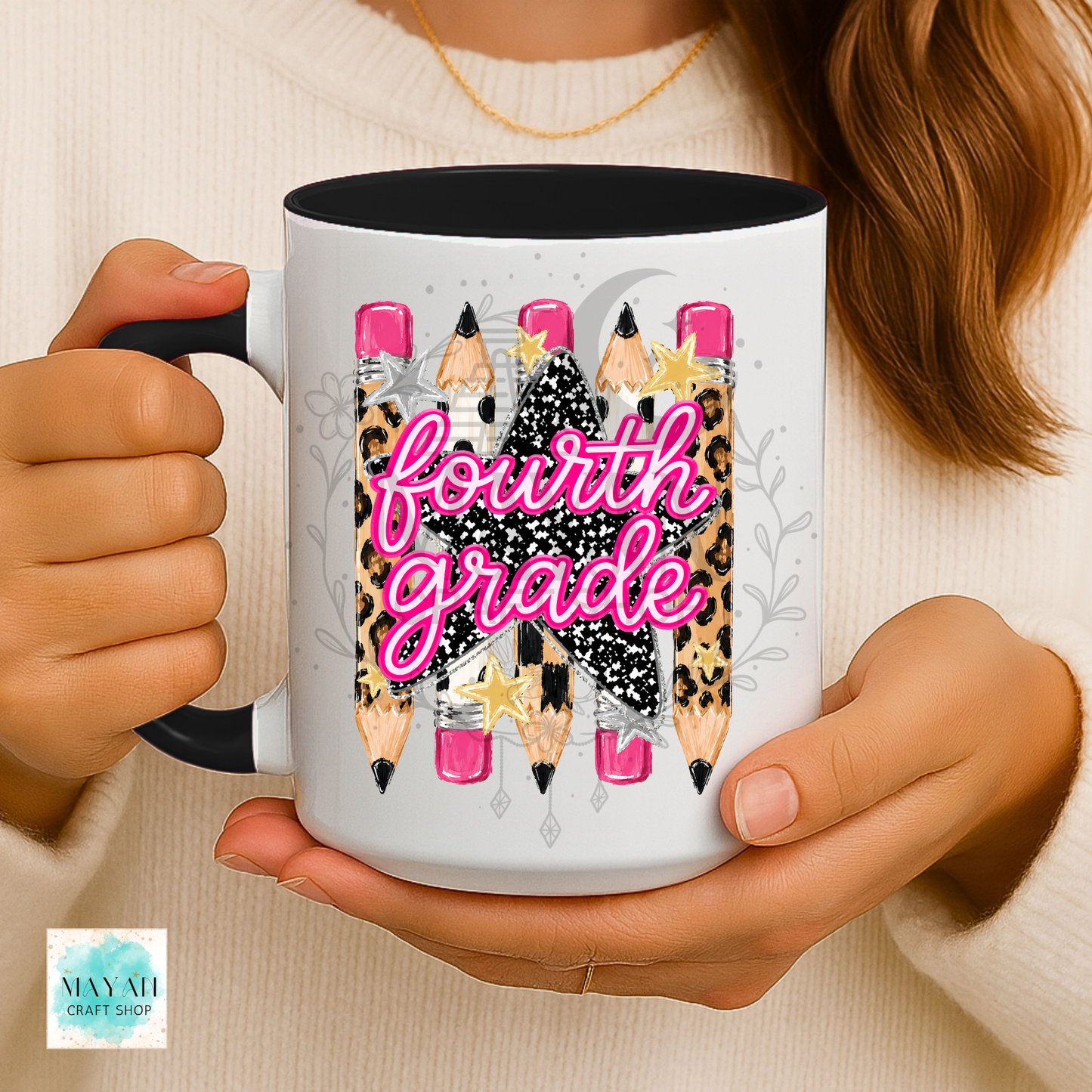 A person holding a white ceramic mug with a black handle and interior. The mug features a graphic design of pencils with leopard print, black glitter patterns, pink erasers, and the words 'fourth grade' in bright pink script.