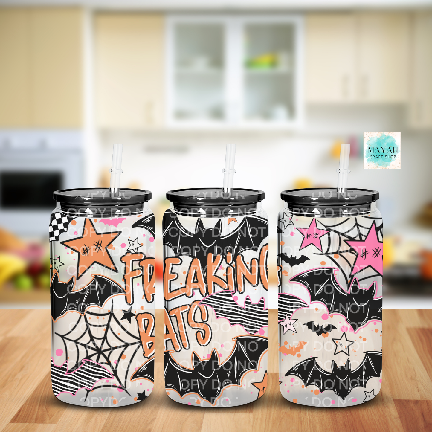 Three tall insulated tumblers with black lids and straws, decorated with a Halloween-themed bat pattern in black, white, pink, orange, and gray, set on a wooden table with a blurred kitchen background.