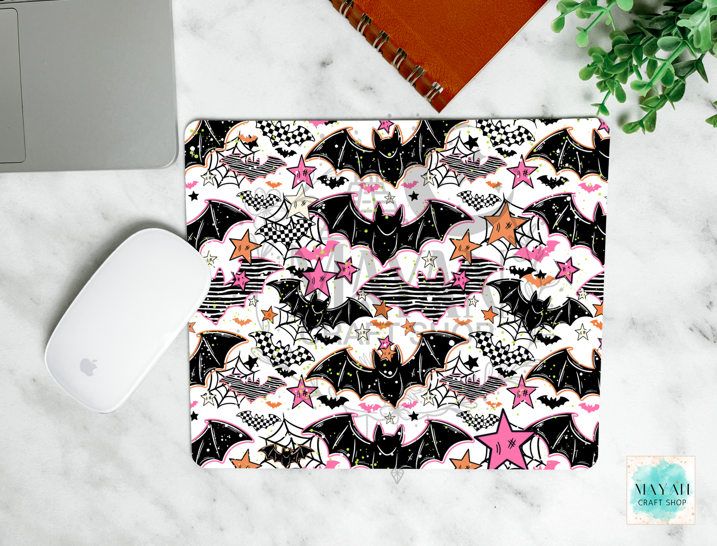 A mouse pad with a Halloween-themed pattern featuring black bats, pink stars, white clouds, and spiderwebs, shown in a flat-lay arrangement on a white marble surface next to a white computer mouse, a silver laptop corner, an orange spiral notebook, and a green plant.