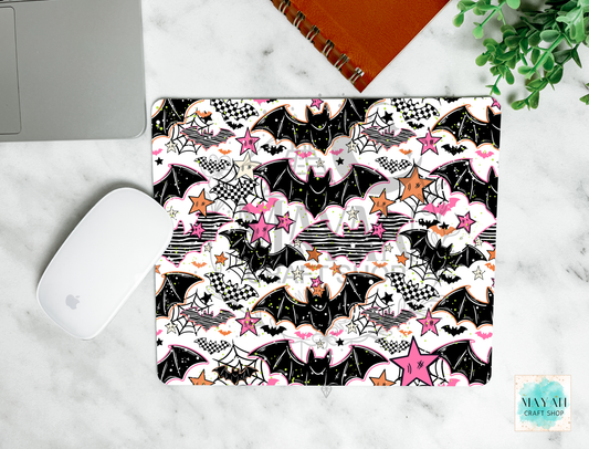 A mouse pad with a Halloween-themed pattern featuring black bats, pink stars, white clouds, and spiderwebs, shown in a flat-lay arrangement on a white marble surface next to a white computer mouse, a silver laptop corner, an orange spiral notebook, and a green plant.