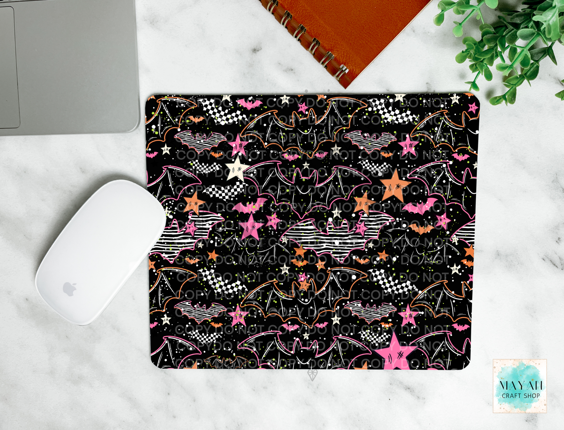 A mouse pad with a black background and a colorful pattern of bats, stars, checkered flags, and lightning bolts, shown in a flat-lay setting on a white marble surface with a laptop mouse, notebook, and plant.