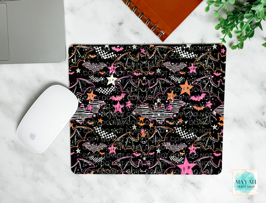 A mouse pad with a black background and a colorful pattern of bats, stars, checkered flags, and lightning bolts, shown in a flat-lay setting on a white marble surface with a laptop mouse, notebook, and plant.