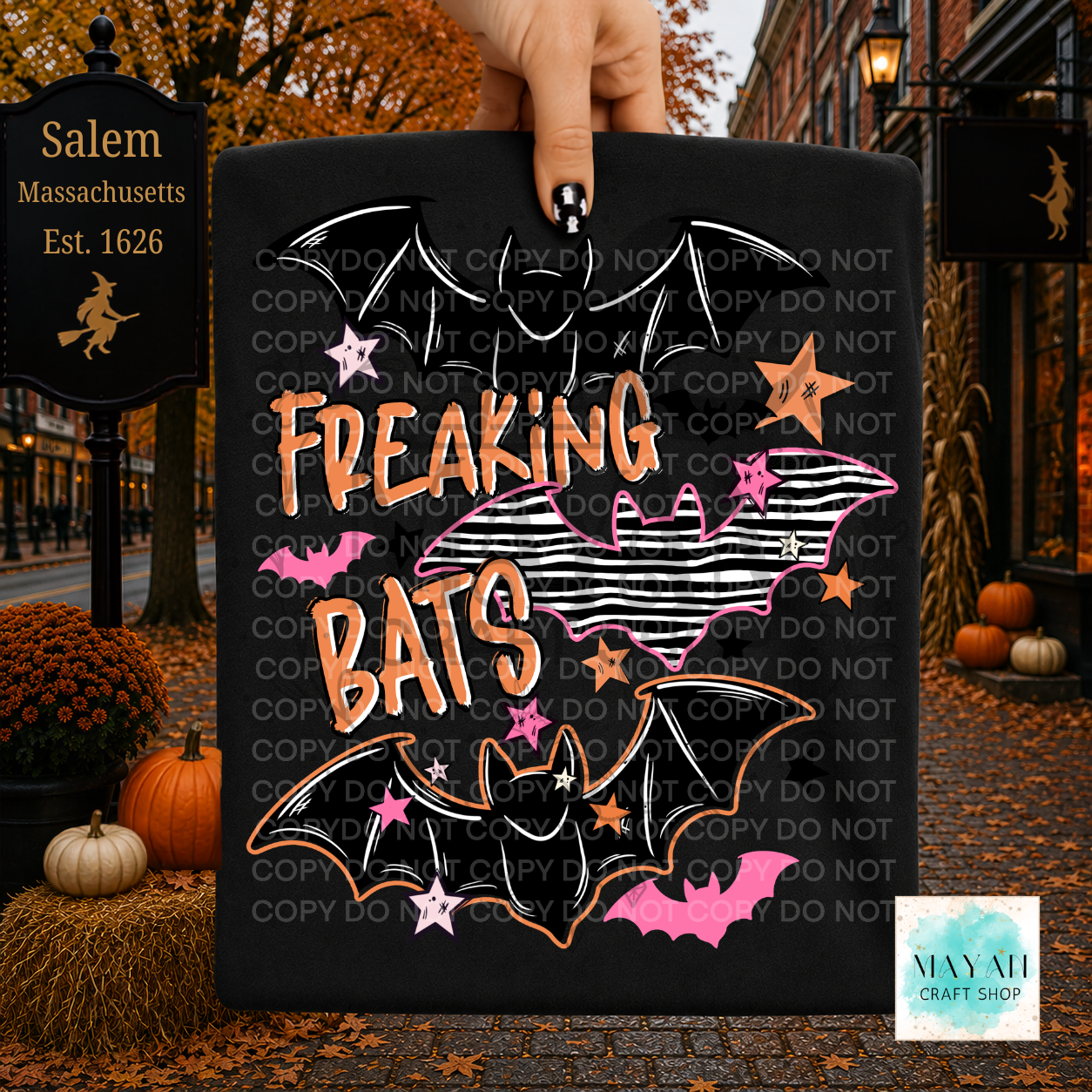 A hand holds a black rectangular sign featuring a Halloween design with the words “FREAKING BATS,” illustrated bats, pink stars, and a striped bat shape. The background shows an autumn street scene with pumpkins, fall leaves, and a sign.
